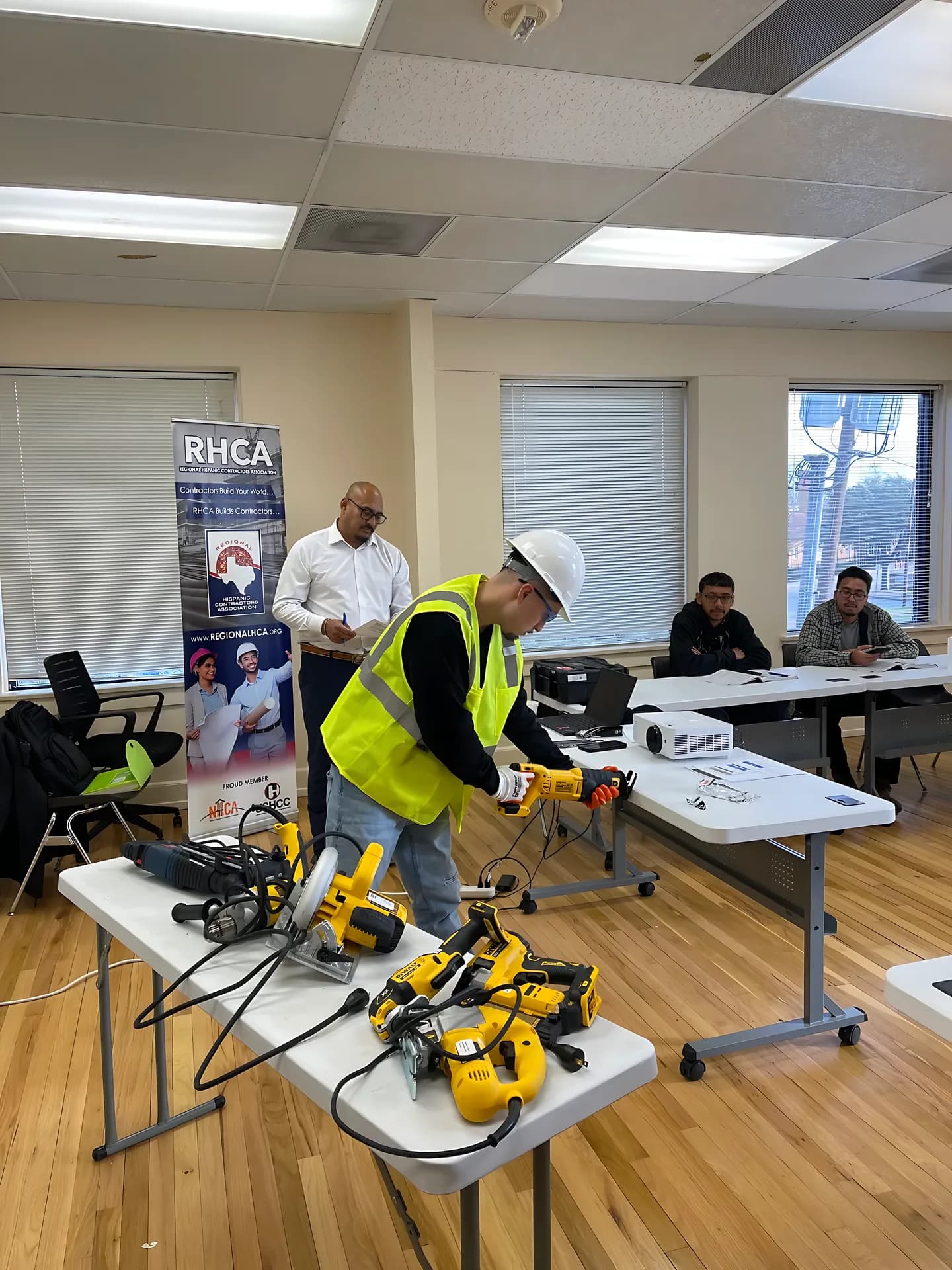 A participant practices with a power tool while an instructor and others observe in a classroom.